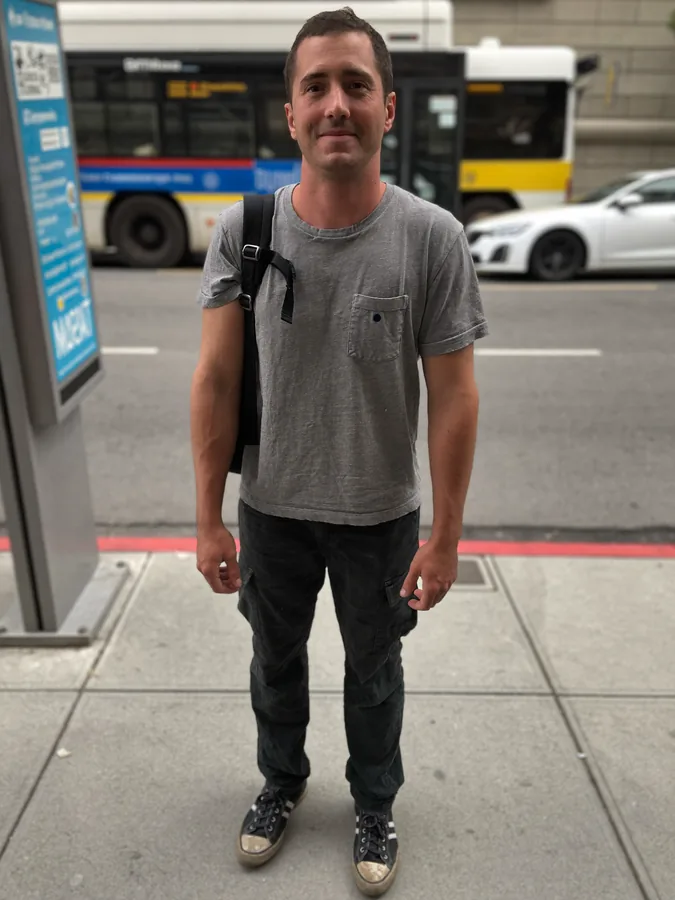 A man at a bus stop in a grey t-shirt and dark trousers with a backpack